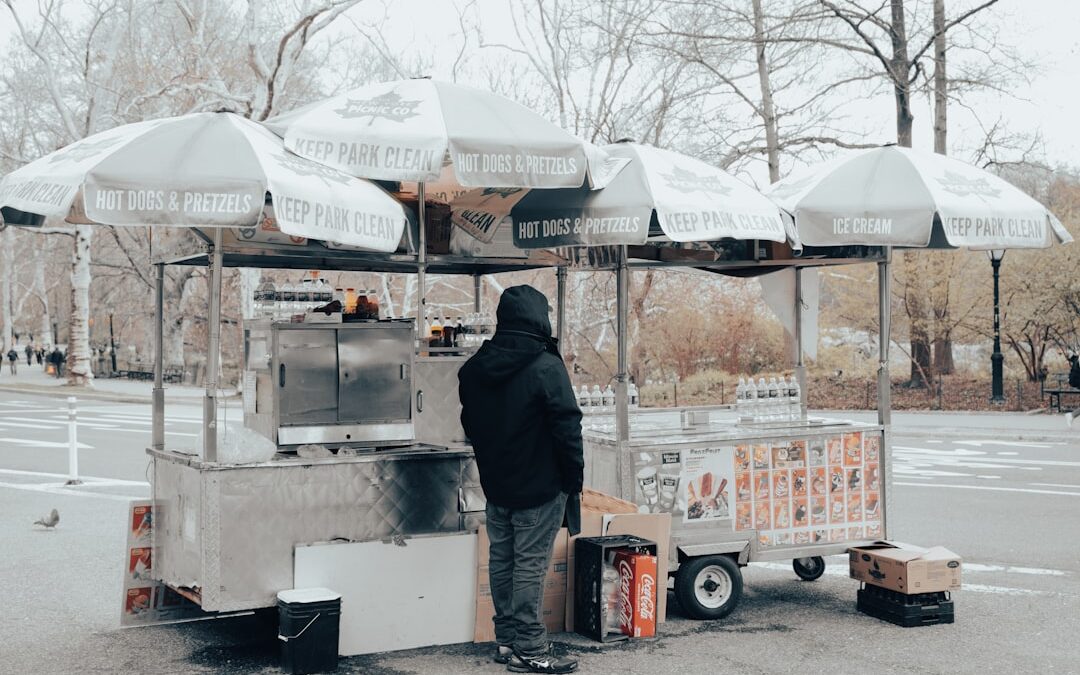 Food cart with umbrellas displaying "Hot Dogs & Pretzels" and "Ice Cream," featuring a vendor in a black coat, set in a park environment.