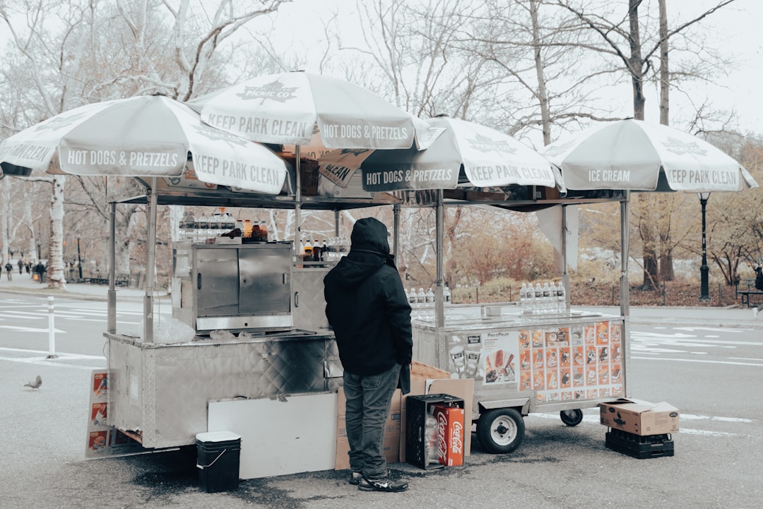 Food pickup cart in a park, featuring umbrellas labeled "Hot Dogs & Pretzels" and "Ice Cream," with a figure in a black jacket standing nearby, highlighting convenient dining options.