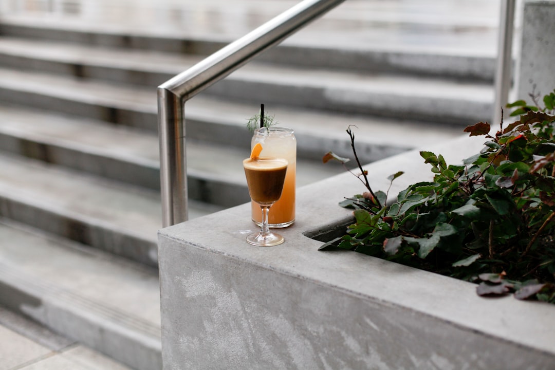 Two stylish cocktails rest on a concrete ledge beside a stairway — one dark espresso martini in a coupe glass and one light orange cocktail in a tall glass garnished with herbs, creating a modern and minimal outdoor setting.